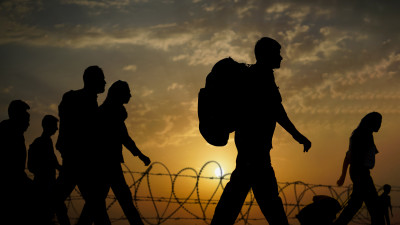 Migrants walking at sunset with barbed wire in foreground.