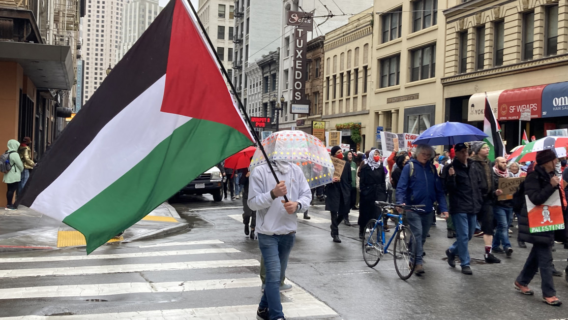 A demonstrator carries a large Palestinian flag and an umbrella while marching against a possible Israeli invasion of Rafah. [Brooke Anderson/The New Arab]